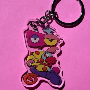 May include: A clear acrylic keychain featuring a colorful cartoon character with a pink head, yellow eye, and a blue lightning bolt. The character has a yellow and red body, purple pants, and a large silver key ring.