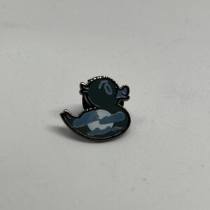 May include: A black enamel pin of a cartoon duck with a moon and clouds inside. The duck is facing right and has a white beak and eyes.