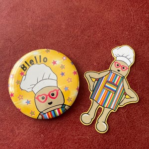 May include: A yellow badge with the word "Blello" and a cartoon chef wearing a white hat and pink glasses. A matching die-cut sticker of the chef in a striped apron is also present.