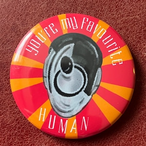 May include: A circular badge with a red base and orange and yellow rays. It features a black and white illustration of a person's head and the white text "you're my favourite human".
