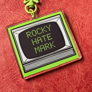 May include: A square acrylic keychain featuring a lime green border and a star-shaped clasp. The center displays a retro television graphic with a black screen containing the pixelated white text ROCKY HATE MARK in three stacked lines.