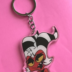 May include: A clear acrylic keychain featuring a cartoon character with red skin, yellow eyes, and a black and white striped hairstyle. The character has a red bow tie and is attached to a silver keyring.