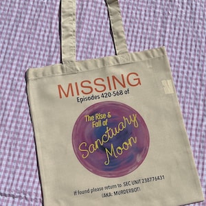 May include: A beige canvas tote bag with the word "MISSING" in red. The bag has text about "The Rise & Fall of Sanctuary Moon" and a return address. The bag has two beige handles.
