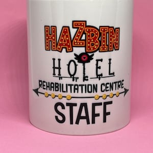 May include: White ceramic mug with a black and red graphic design that says "Hazbin Hotel Rehabilitation Centre Staff". The graphic design features a red eye with a white heart in the center.