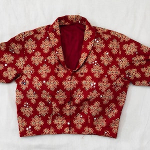 Può includere: Una camicetta rossa con un motivo floreale dorato ripetuto e piccoli accenti a specchio. La camicetta ha un colletto e maniche corte. Il tessuto sembra leggero.