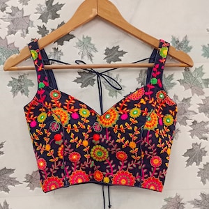 May include: A navy blue embroidered top with a V-neckline and spaghetti straps. The top features a vibrant floral pattern in shades of pink, orange, yellow, and green. The garment is displayed on a wooden hanger.