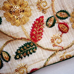 Pu&ograve; includere: Primo piano di un tessuto color crema con ricami dorati, verdi e rossi. Il ricamo presenta motivi floreali con paillettes.
