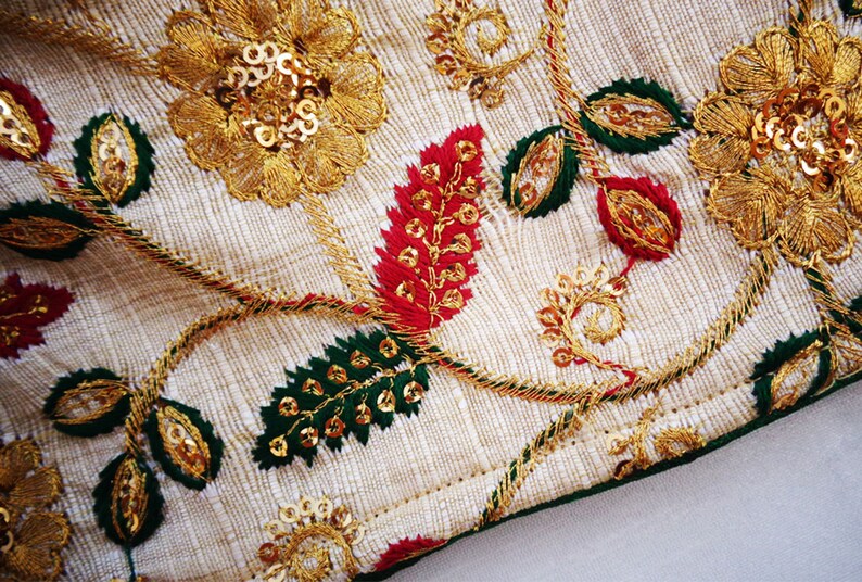 Pu&ograve; includere: Primo piano di un tessuto color crema con ricami dorati, verdi e rossi. Il ricamo presenta motivi floreali con paillettes.