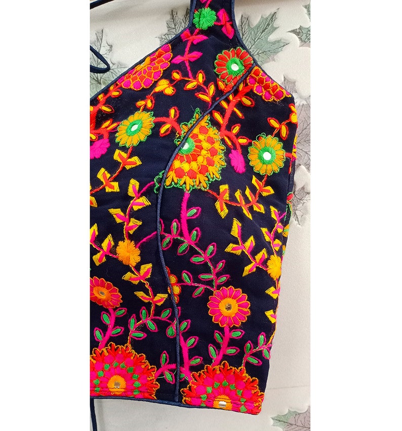 May include: A navy blue halter top with vibrant floral embroidery. The embroidery features pink, yellow, orange, and green flowers and leaves. The top has a simple design with a halter neck and a straight hemline.