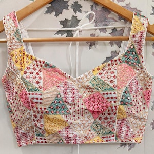 以下が含まれることがあります： Vネックの白いノースリーブトップス。ピンク、イエロー、グリーン、レッドの刺繍が施されたパッチワークデザイン。木製のハンガーにかかっています。