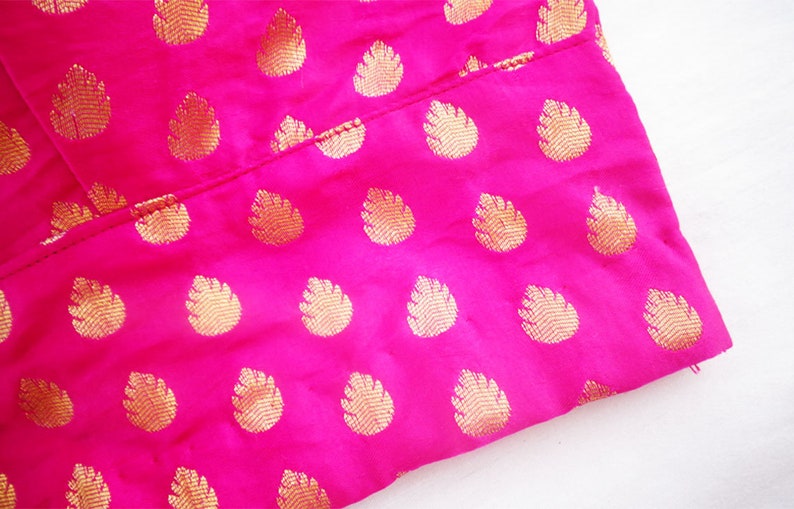 May include: Close-up of a vibrant pink fabric with a repeating gold leaf pattern. The fabric is folded, revealing the texture and sheen of the material. The gold pattern adds a touch of elegance to the bright pink background.