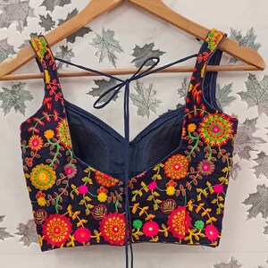 May include: A navy blue embroidered top with a floral design in shades of orange, pink, yellow, and green. The top has a scoop neckline and tie-up straps. The garment is displayed on a wooden hanger.