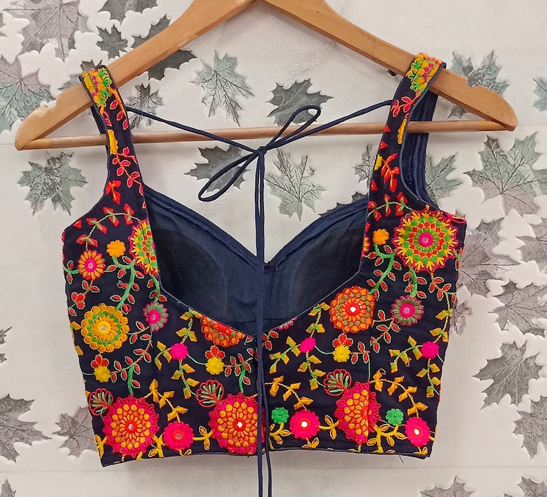 May include: A navy blue embroidered top with a floral design in shades of orange, pink, yellow, and green. The top has a scoop neckline and tie-up straps. The garment is displayed on a wooden hanger.