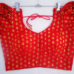 May include: A red blouse with gold leaf-shaped patterns. The blouse has short, puffed sleeves and a low, rounded neckline. A red tie is at the back of the neck. The fabric has a slight sheen, and the blouse is hanging on a hanger.