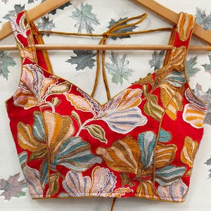 Könnte beinhalten: Ein rotes, besticktes Oberteil mit V-Ausschnitt und breiten Trägern. Das florale Design weist Gold-, Orange-, Blau- und Rosatöne auf. Das Oberteil hängt an einem Holzbügel mit Krawattenverschluss.