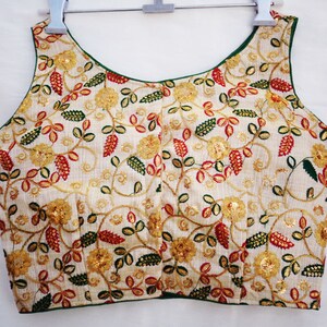Pu&ograve; includere: Una blusa bianca senza maniche con scollo verde e ricamo floreale in tonalit&agrave; rosso, verde e oro.