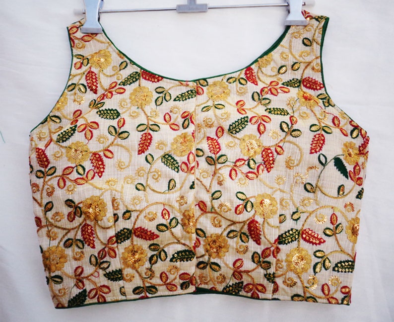 Pu&ograve; includere: Una blusa bianca senza maniche con scollo verde e ricamo floreale in tonalit&agrave; rosso, verde e oro.