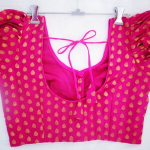May include: A bright pink blouse with gold leaf-shaped patterns. The blouse features short, puffed sleeves and a scoop neckline. A tie closure is visible at the back. The garment is hanging on a silver hanger.