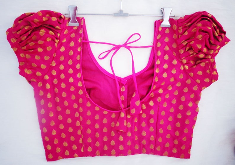 May include: A bright pink blouse with gold leaf-shaped patterns. The blouse features short, puffed sleeves and a scoop neckline. A tie closure is visible at the back. The garment is hanging on a silver hanger.