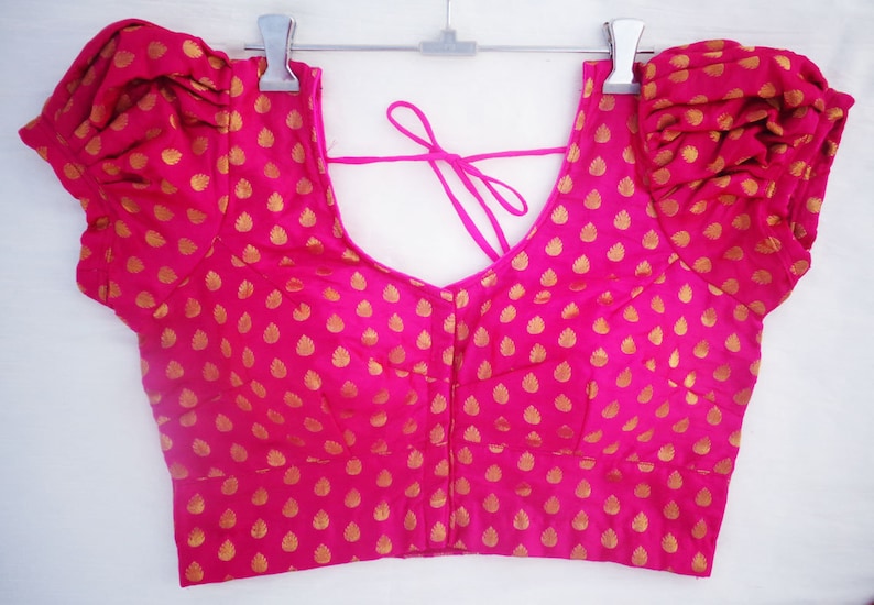 May include: A hot pink blouse with short, puffed sleeves and a low V-neck. The fabric is patterned with small, gold-colored leaf shapes. A pink tie is visible at the back of the neck. The blouse is hanging on a silver hanger.