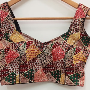 以下が含まれることがあります： 赤、緑、黄、金の色合いの幾何学模様が刺繍された、黒いノースリーブのブラウス。ブラウスは、スパンコールとビーズで飾られています。