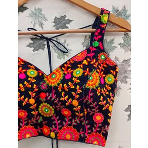 May include: A black embroidered top with a V-neckline and tie straps. The top is adorned with vibrant floral embroidery in shades of pink, yellow, orange, and green. The straps and neckline are outlined in a dark blue color.