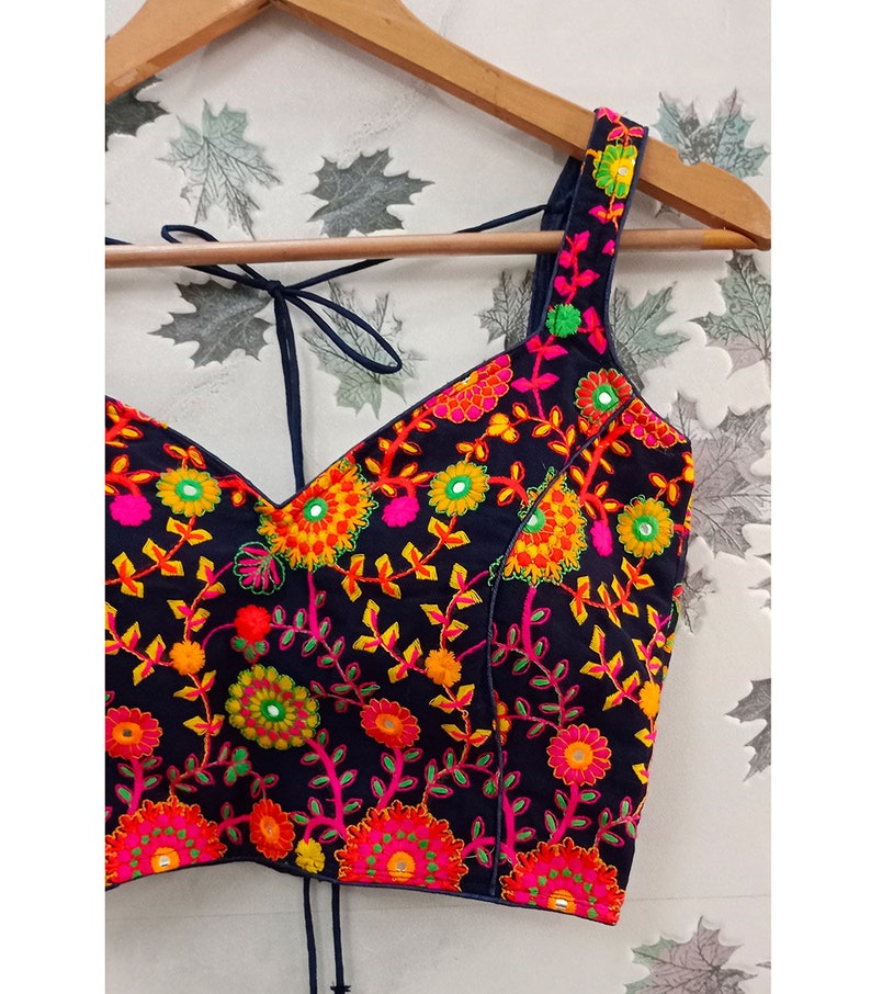 May include: A black embroidered top with a V-neckline and tie straps. The top is adorned with vibrant floral embroidery in shades of pink, yellow, orange, and green. The straps and neckline are outlined in a dark blue color.