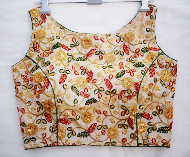 Pu&ograve; includere: Una blusa bianca senza maniche con un motivo floreale ricamato in filo dorato, verde e rosso. La blusa ha una scollatura rotonda e un bordo verde.