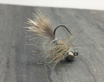 May include: A close-up of a fly fishing lure. The lure features a gray, fluffy body with iridescent accents and a dark metal hook and bead. The fly is designed to mimic an insect.