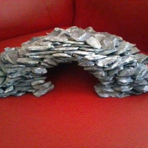 Peut inclure: Un arche en pierre grise, construite à partir de morceaux d'ardoise superposés. L'arche a un sommet arrondi et un centre creux, adapté à la décoration de terrariums ou d'aquariums. La structure est posée sur un fond rouge.