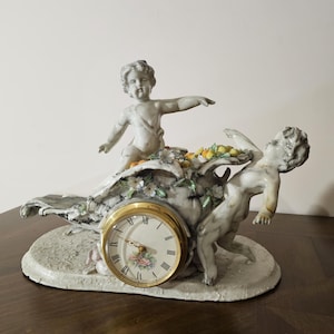 Puede incluir: Un reloj de porcelana blanca y gris con dos figuras de querubines. La esfera del reloj es dorada con números romanos negros y un diseño floral. La base es de forma ovalada, con detalles florales y de hojas.