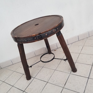 Puede incluir: Un taburete redondo de madera con un anillo de metal alrededor de la parte superior y tres patas. El taburete tiene un anillo de metal en la parte inferior para soporte.