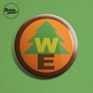 May include: A brown and orange button featuring a green pine tree with the letters "W" and "E" inside. The button is circular and has a glossy finish.