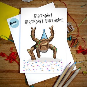 May include: A birthday card with the word "BIRTHDAY!" repeated three times. The card features a cartoon creature wearing a party hat, with confetti at the bottom. Pencils and other stationery are visible.