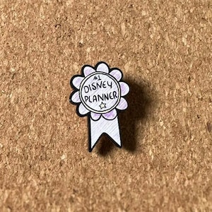May include: A white and purple Disney Planner award pin with a black outline. The pin features the text "#1 Disney Planner" in a circular design, with a star at the bottom. The pin is on a brown corkboard surface.