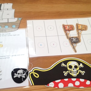 May include: A set of pirate-themed educational games for kids. The set includes a pirate ship, a pirate hat, an eye patch, and a game board with numbered circles and pirate-themed images. The game board has the text "Boat Treasure Number Match".