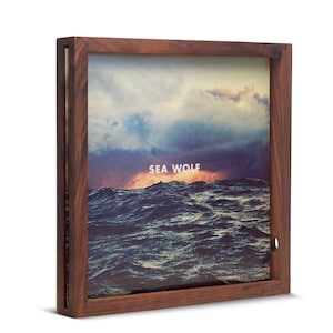May include: A wooden frame with a record album inside. The album cover features a photograph of a stormy sea with a sunset in the background. The album title "SEA WOLF" is printed in white on the cover.