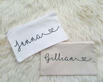 May include: Two white canvas zippered pouches with black lettering. The top pouch has the name "Jenna" with a heart symbol. The bottom pouch has the name "Gillian" with a heart symbol.
