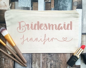 May include: A white canvas makeup bag with a zipper closure. The bag has the words "Bridesmaid Jennifer" printed in pink on the front. The bag is lying on a wooden surface with makeup brushes and a red lipstick.