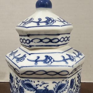 Peut inclure: Pot en céramique blanc et bleu avec couvercle. Le pot est de forme hexagonale et décoré de motifs floraux bleus. Le couvercle est surmonté d'un bouton bleu. Le pot est probablement destiné au stockage ou à la décoration.