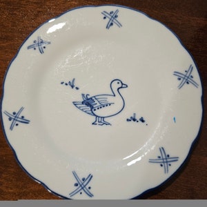 May include: A white ceramic plate with a blue rim and a blue and white illustration of a duck. The plate has a decorative blue pattern of criss-crossed lines around the edge.