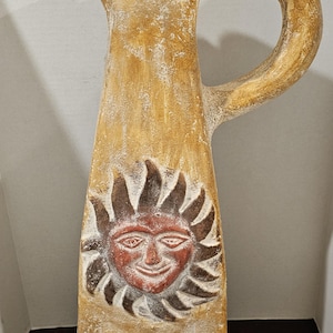 May include: Tan ceramic pitcher with a handle and spout. The front features a carved sun face design in red and brown. The word "MEXICO" is inscribed at the base. A decorative piece.