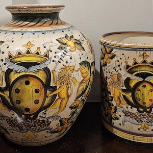 May include: Two ceramic decorative vases. The larger vase is round with a narrow neck, and the smaller one is cylindrical. Both are cream-colored with a repeating design of lions, crowns, and shields in yellow, orange, and blue.