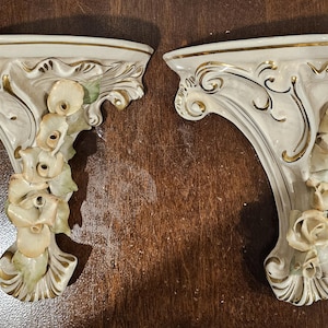 May include: Two ornate, off-white wall shelves with gold trim and floral accents. The shelves have a curved design with decorative carvings and are likely made of ceramic or porcelain. A classic home decor item.