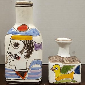 May include: Two ceramic decorative bottles. One is tall and rectangular, featuring a painted face with a mustache, a red spot, and blue and purple accents. The other is a small, square bottle with a yellow bird design.