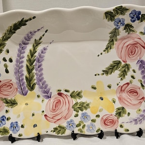 May include: A rectangular, white ceramic serving platter with a scalloped edge. The platter is hand-painted with pink roses, blue and yellow flowers, and lavender sprigs. The platter is supported by a black metal stand.