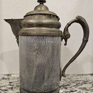 May include: A silver and gray metal pitcher with a decorative handle and a textured surface.