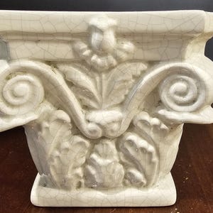 May include: White ceramic planter shaped like a column capital with intricate scrollwork and floral details. The planter has a crackled glaze finish.