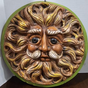 May include: A round, sculpted wall hanging featuring a face with a long beard and mustache. The face is brown with black eyes and white accents. The beard and hair are a mix of yellow and brown. The outer edge is green.
