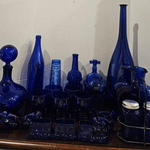 May include: A collection of cobalt blue glass bottles, decanters, glasses, and serving pieces. The assortment includes a tall, slender bottle, a decanter with a round stopper, and a set of salt and pepper shakers.